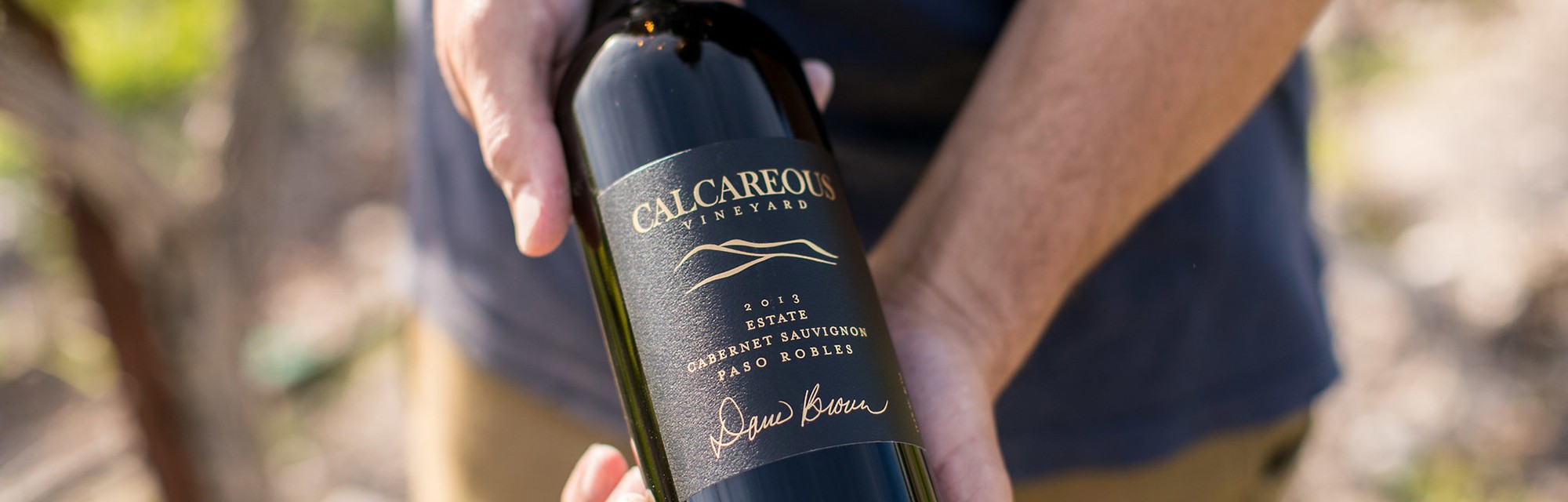 Winemaker Jason Joyce stands in the vineyard holding a bottle of 2013 Signature Cabernet Sauvignon